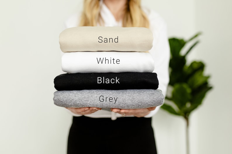 K&ouml;nnte beinhalten: Ein Stapel gefalteter T-Shirts in verschiedenen Farben: Sand, Wei&szlig;, Schwarz und Grau.