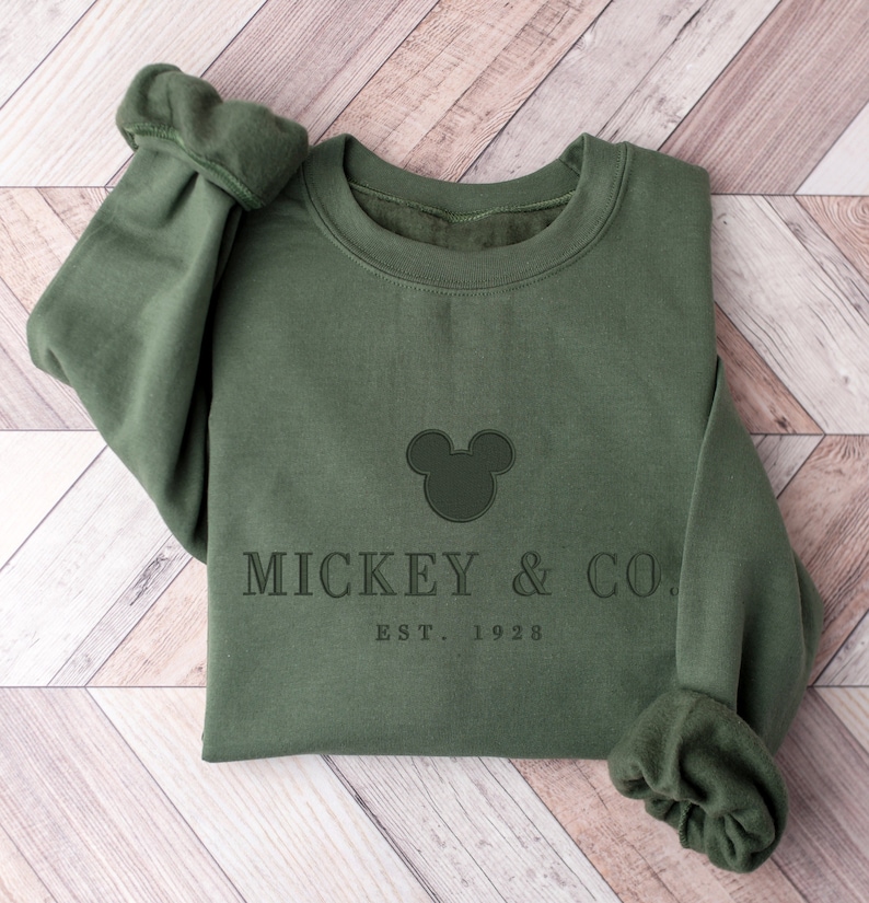 Op de afbeelding: Een olijfgroene sweater met ronde hals en een donkergroen geborduurde Mickey Mouse-hoofdsilhouet, gecentreerd boven de tekst MICKEY & CO. en de kleinere tekst EST. 1928. Het kledingstuk heeft lange mouwen met geribbelde boorden en een geribbelde hals.