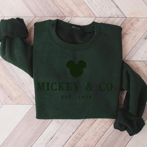 Op de afbeelding: Een donker bosgroene sweater met ronde hals en een zwart geborduurd Mickey Mouse-oorsilhouet, gecentreerd boven de tekst MICKEY & CO. en de kleinere tekst EST. 1928, gedrukt in een strak, donker schreefloos lettertype op de borst.