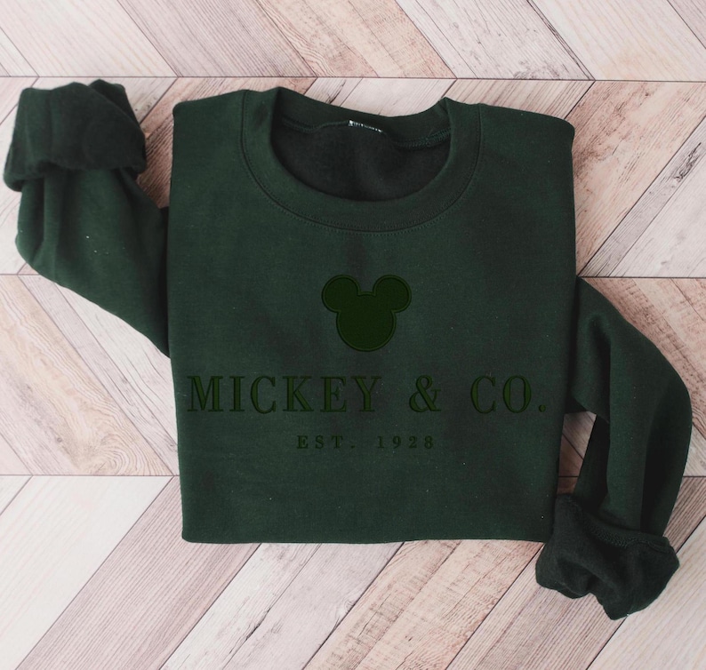 Op de afbeelding: Een donker bosgroene sweater met ronde hals en een zwart geborduurd Mickey Mouse-oorsilhouet, gecentreerd boven de tekst MICKEY & CO. en de kleinere tekst EST. 1928, gedrukt in een strak, donker schreefloos lettertype op de borst.