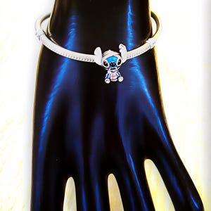 May include: A silver-toned bracelet featuring a charm of the character Stitch from the animated film Lilo & Stitch. The charm is blue, white, and red, with a detailed design. The bracelet is on a black surface.