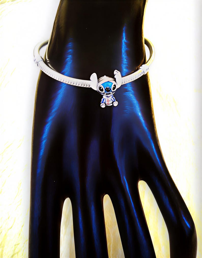 May include: A silver-toned bracelet featuring a charm of the character Stitch from the animated film Lilo & Stitch. The charm is blue, white, and red, with a detailed design. The bracelet is on a black surface.