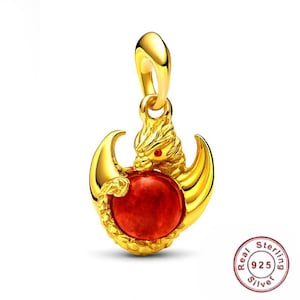 May include: A gold-colored pendant featuring a dragon design with a red gemstone center. The dragon has detailed features and is holding the gemstone. The pendant has a loop for a chain.