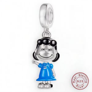 Może przedstawiać: Srebrny charms przedstawiający postać z kreskówki z czarnymi włosami i niebieską sukienką. Charms jest zawieszony na srebrnej pętli i ma stempel srebra 925. Postać się uśmiecha.