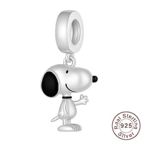 Könnte beinhalten: Ein silberner Charm-Anhänger mit Snoopy, dem Zeichentrickhund, mit schwarzem Ohr und Nase und einem lächelnden Gesicht. Der Charm hat eine Schlaufe zum Befestigen an einem Armband oder einer Halskette. Der Charm ist aus Sterlingsilber.