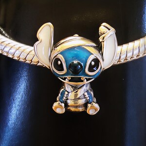 May include: A charm featuring the character Stitch from Lilo & Stitch. The charm is blue, silver, and white, with a detailed design. It is attached to a silver bracelet with a snake chain design. The charm is approximately 1 inch tall.
