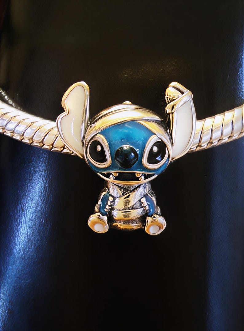May include: A charm featuring the character Stitch from Lilo & Stitch. The charm is blue, silver, and white, with a detailed design. It is attached to a silver bracelet with a snake chain design. The charm is approximately 1 inch tall.