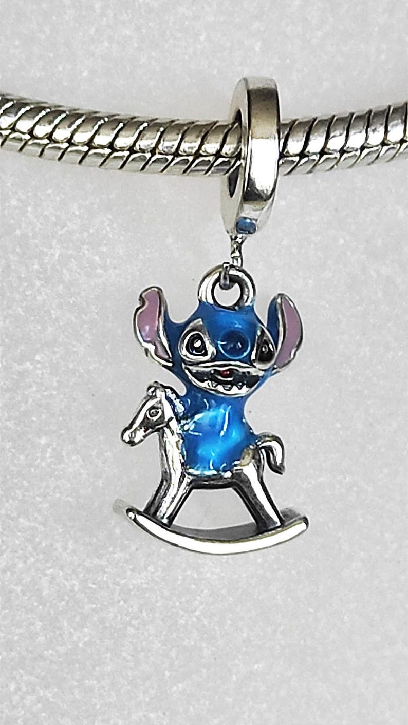 Pode incluir: Um berloque azul e prateado com um personagem de desenho animado em um cavalo de balan&ccedil;o. O berloque tem um corpo azul, orelhas rosa e uma base de cavalo de balan&ccedil;o prateada. O berloque &eacute; preso a uma pulseira de prata.