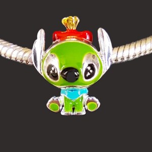 May include: A green and silver charm depicting a cartoon character with large black eyes, a red bow, and a gold crown. The charm has a blue collar and is attached to a silver chain. A collectible item.