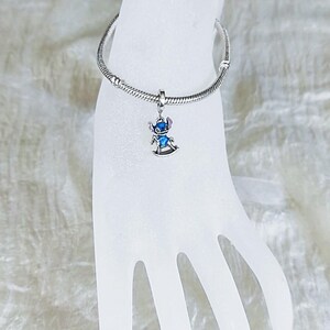 Pode incluir: Uma pulseira de prata com um berloque com um personagem de desenho animado azul e prateado em um cavalo de balan&ccedil;o. A pulseira &eacute; exibida em uma m&atilde;o branca. O berloque &eacute; detalhado.