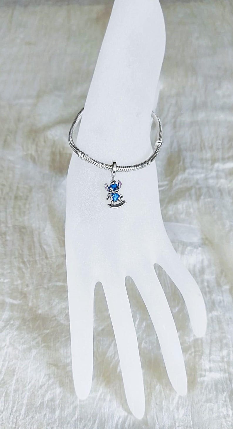 Pode incluir: Uma pulseira de prata com um berloque com um personagem de desenho animado azul e prateado em um cavalo de balan&ccedil;o. A pulseira &eacute; exibida em uma m&atilde;o branca. O berloque &eacute; detalhado.