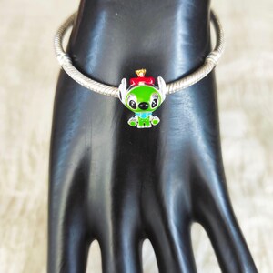May include: A silver charm bracelet with a green, red, and blue Stitch charm. The charm features Stitch wearing a red bow and a gold crown. The bracelet is displayed on a black hand.