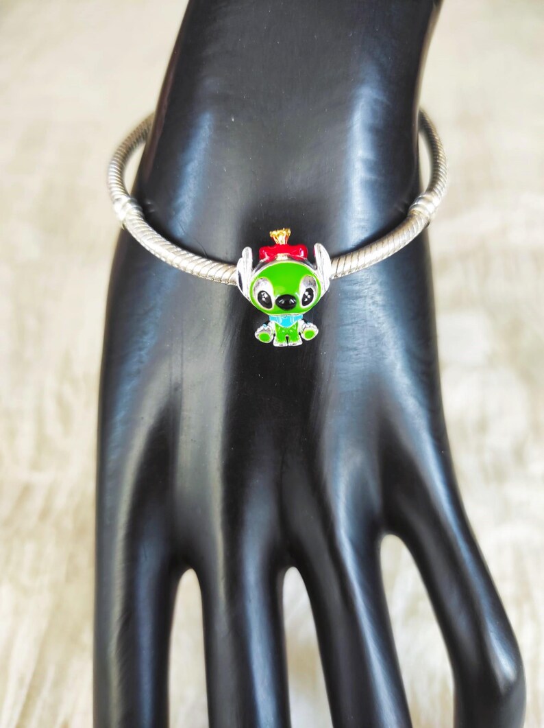 May include: A silver charm bracelet with a green, red, and blue Stitch charm. The charm features Stitch wearing a red bow and a gold crown. The bracelet is displayed on a black hand.