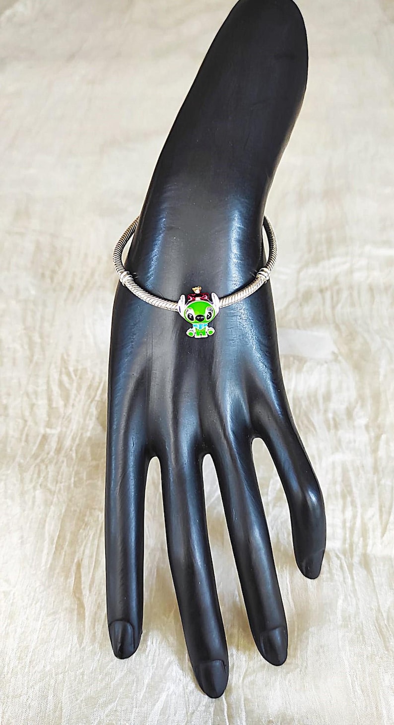 May include: A silver charm bracelet with a colourful Stitch charm. The charm features a green, red, and white design of the character. The bracelet is displayed on a black hand-shaped jewellery display.