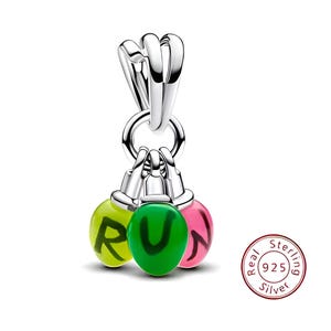 Puede incluir: Un charm de plata de ley con tres esferas de colores. Cada esfera es amarilla, verde o rosa, con la palabra "RUN" impresa. El charm está diseñado para ser adjuntado a una pulsera o collar.