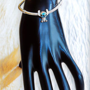 May include: A silver bracelet with a charm of the character Stitch from Lilo & Stitch. The charm is blue and white, and the bracelet is on a black hand-shaped display.