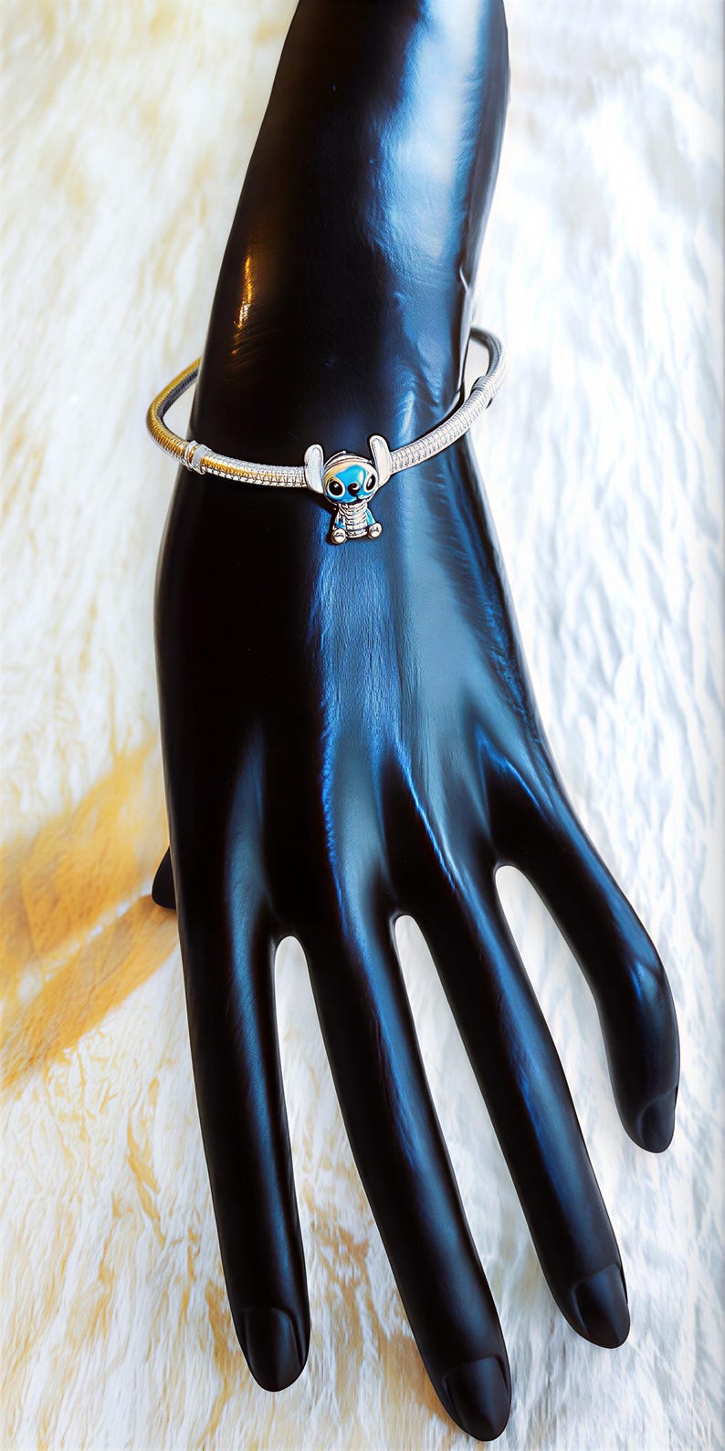 May include: A silver bracelet with a charm of the character Stitch from Lilo & Stitch. The charm is blue and white, and the bracelet is on a black hand-shaped display.