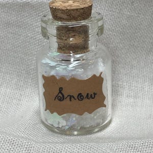 May include: A small, clear glass bottle with a cork stopper, filled with iridescent, glittery material. A brown paper label on the bottle reads "Snow" in handwritten script. The bottle is set against a neutral background.