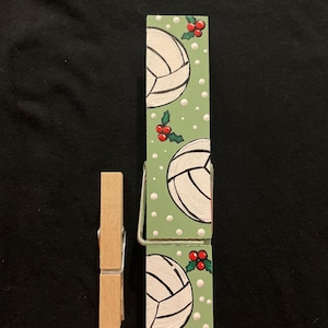 May include: Two wooden clothespins. One is a standard clothespin, the other is painted green with white volleyballs, red berries, and white dots. The volleyball-themed clothespin is much larger than the standard clothespin.