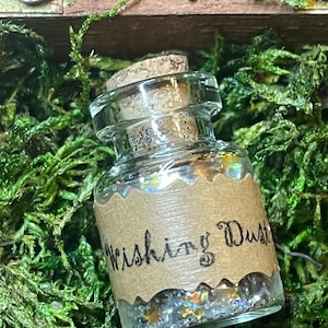 May include: A small glass bottle with a cork stopper, labeled "Wishing Dust." The bottle is filled with iridescent glitter and sits amidst green moss. The label is a light brown color with the words "Wishing Dust" in black script.