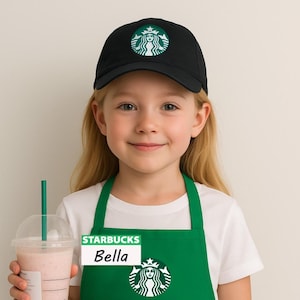 May include: A black baseball cap with a white and green Starbucks logo, a green apron featuring a white Starbucks logo and a name tag that reads Starbucks Bella, and a clear plastic cup containing a light pink beverage with a green straw.