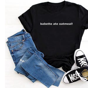 May include: A black t-shirt with the text "babette ate oatmeal!" in white, paired with blue jeans and black canvas sneakers with white laces and soles. The jeans are folded and have a distressed look.