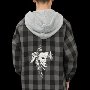 May include: A gray and black plaid flannel shirt with a gray hood. The back of the shirt features a white graphic of a character holding a knife. The shirt is buttoned and has long sleeves.