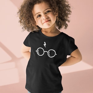 May include: A young girl wearing a black t-shirt with a white lightning bolt and glasses graphic. The girl has curly brown hair and is smiling.