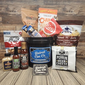 May include: A black bucket filled with various snacks, including popcorn, pretzels, hot sauce, mustard, and a bar mix. The bucket has a blue and white label that says "Detroit Can Co.".