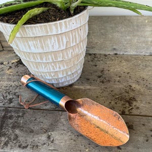 May include: A small copper gardening trowel with a teal blue handle and a leather strap. The trowel has a hammered texture and is resting on a wooden surface.