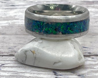 May include: A silver ring with a green and blue opal inlay. The ring is sitting on a white rock.