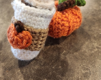 May include: Two crocheted fall-themed decorations: a coffee cup with whipped cream and a pumpkin. The coffee cup is brown, white, and orange, and the pumpkin is orange with a green stem.
