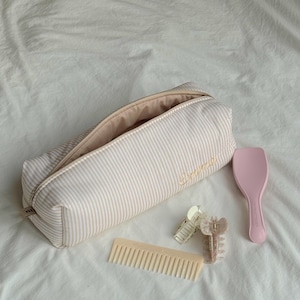 May include: A beige and white striped pencil case embroidered with the name "Emma" in gold, a pink hairbrush, two hair clips, and a beige comb are arranged on a white bedsheet.