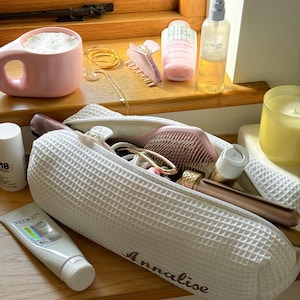 May include: A collection of beauty and self-care items arranged on a wooden surface. Items include a pink mug, hair clips, jewelry, a personalized white waffle-weave cosmetic bag, hairbrushes, skincare products, and a candle.