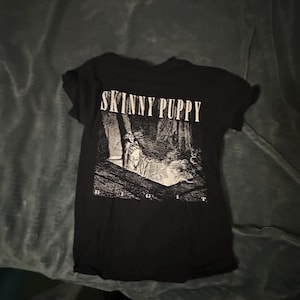 May include: A black short-sleeve t-shirt featuring the white text SKINNY PUPPY at the top and the word DIGIT at the bottom. The center graphic depicts a black and white illustration of a figure standing within a dark, cavernous architectural space.