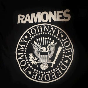 Puede incluir: Una camiseta negra con el texto blanco RAMONES en la parte superior. Debajo hay un gráfico circular que se asemeja a un sello presidencial con los nombres Johnny, Joey y Dee Dee, y un águila que sostiene flechas y un bate de béisbol con el texto LOOK OUT BELOW.