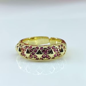 May include: A gold-colored ring with a band adorned with small, triangular-shaped accents and embedded with numerous small, red gemstones. The ring is open-ended, allowing for size adjustment. The ring is displayed on a white surface.