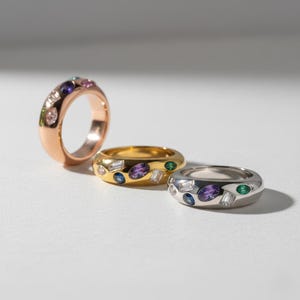 以下が含まれることがあります： ローズゴールド、ゴールド、シルバーの3つの宝石リング。各リングには、紫、緑、白の石など、さまざまな色の宝石が使用されています。リングは白い表面に表示されています。