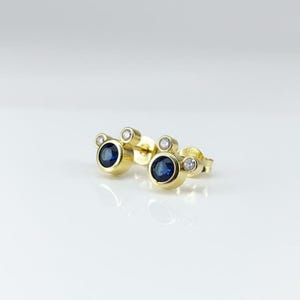 May include: A pair of gold stud earrings with a three-circle design. Each earring features a large, round, dark blue sapphire centre stone, flanked by two smaller, clear, round stones. The earrings are set against a white background.