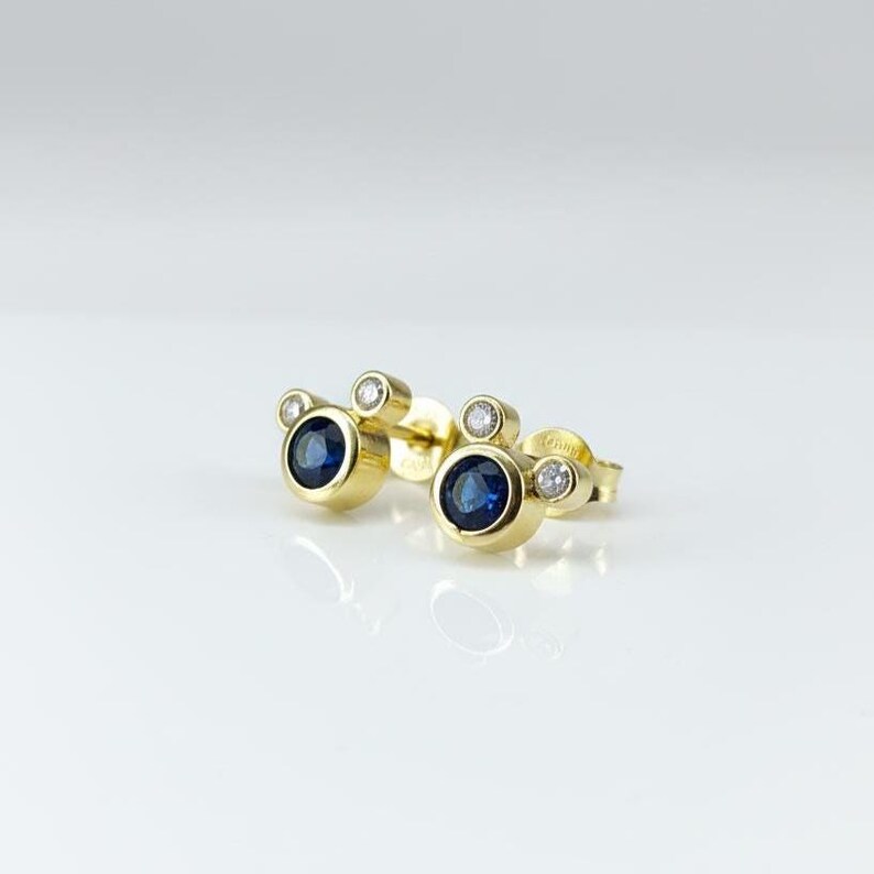 May include: A pair of gold stud earrings with a three-circle design. Each earring features a large, round, dark blue sapphire centre stone, flanked by two smaller, clear, round stones. The earrings are set against a white background.