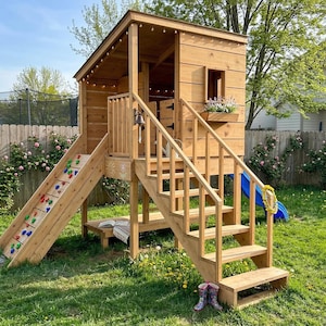 Puede incluir: Una casa de juegos de madera con tobogán, rocódromo y escaleras. La casa de juegos tiene un pequeño alféizar con flores y un tobogán azul. El rocódromo tiene presas de colores. La casa de juegos está en un jardín con césped.