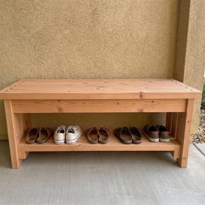 May include: A wooden shoe rack bench with a natural finish. The bench has a top surface and a lower shelf for shoe storage. Several pairs of shoes are visible on the shelf. The bench is made of light-colored wood and is sturdy.