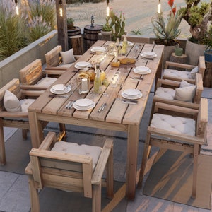 Op de afbeelding: Een houten eettafelset voor buiten met een rechthoekige tafel en zes stoelen. De tafel heeft een natuurlijke houten afwerking en is gedekt met borden, bestek en glazen. De stoelen zijn ook van hout en hebben witte kussens. De set is omgeven door groen en lichtslingers.