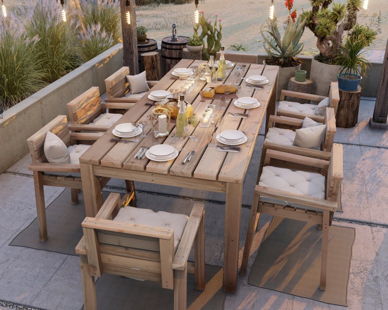 Op de afbeelding: Een houten eettafelset voor buiten met een rechthoekige tafel en zes stoelen. De tafel heeft een natuurlijke houten afwerking en is gedekt met borden, bestek en glazen. De stoelen zijn ook van hout en hebben witte kussens. De set is omgeven door groen en lichtslingers.