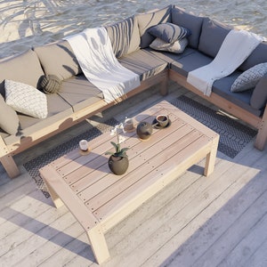May include: A wooden outdoor sectional sofa with gray cushions and white throw blankets. The sofa is set up in a corner with a matching wooden coffee table and two gray rugs. There are two cups of coffee and a teapot on the coffee table.