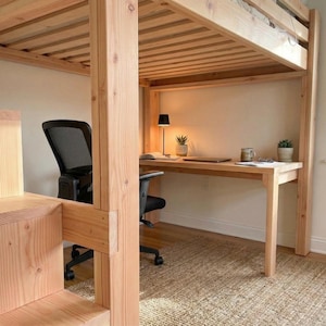 May include: A light-colored wooden loft bed with a built-in desk and stairs. The desk has a black chair, a lamp, and a few items on top. The stairs are on the left side of the bed. The room has a neutral color scheme.