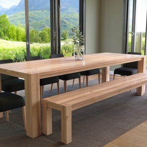 Może przedstawiać: Jasny drewniany stół jadalny z naturalnym wykończeniem i sześcioma krzesłami. Stół jest ustawiony z wazą z zielenią. Do końca stołu przylega pasująca drewniana ławka. Stół i ławka znajdują się w pokoju z dużymi oknami z widokiem na zielony krajobraz.