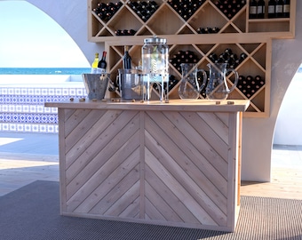 May include: A wooden bar with a chevron pattern on the front. The bar has a glass dispenser with a lid, two glass pitchers, and two ice buckets on the top. There are bottles of wine on shelves behind the bar.