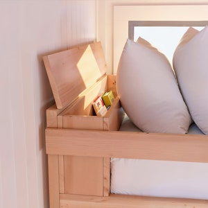May include: A wooden bed frame with a built-in storage compartment. The compartment is open and reveals a variety of items, including books and other small objects. The bed is made up with white bedding and pillows.