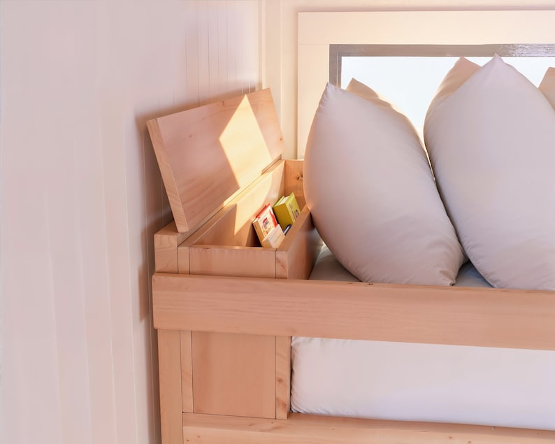 May include: A wooden bed frame with a built-in storage compartment. The compartment is open and reveals a variety of items, including books and other small objects. The bed is made up with white bedding and pillows.
